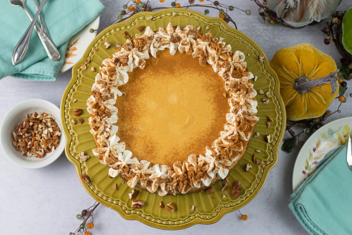 A uncut Pumpkin Cheesecake garnished with whipped cream and chopped pecans on a green glass cake stand.