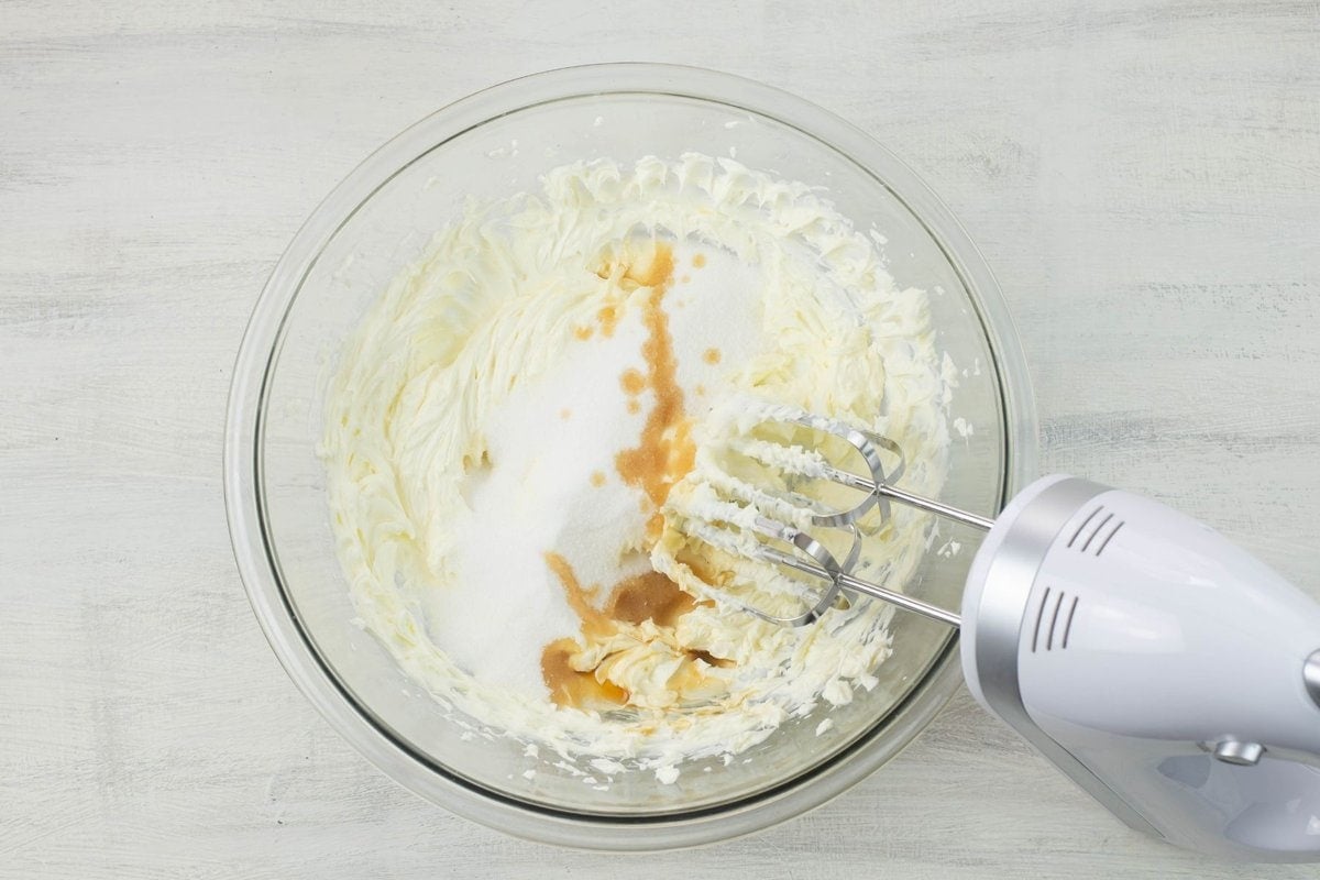 Adding white sugar and vanilla extract to a bowl of beaten cream cheese.