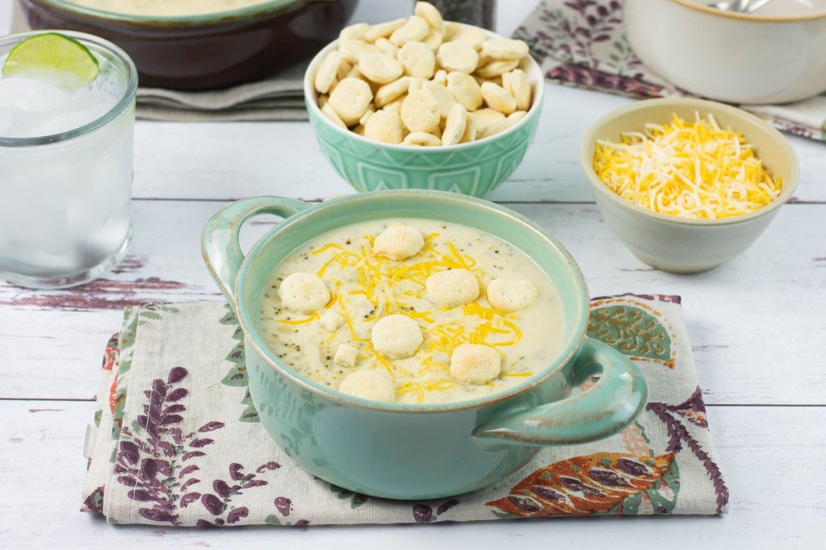 A serving of cream of broccoli soup with a bowl of crackers and shredded cheese on a dinner table.