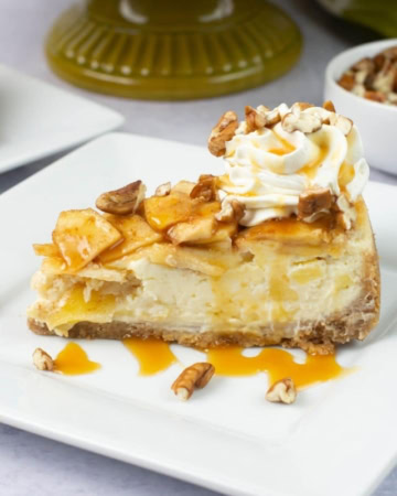 A single slice of apple cheesecake garnished with whip cream, caramel sauce and chopped pecans on a dessert plate.
