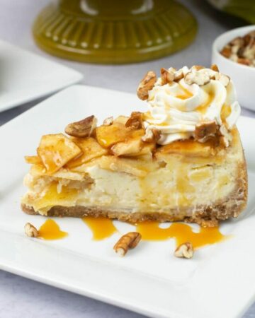 A single slice of apple cheesecake garnished with whip cream, caramel sauce and chopped pecans on a dessert plate.