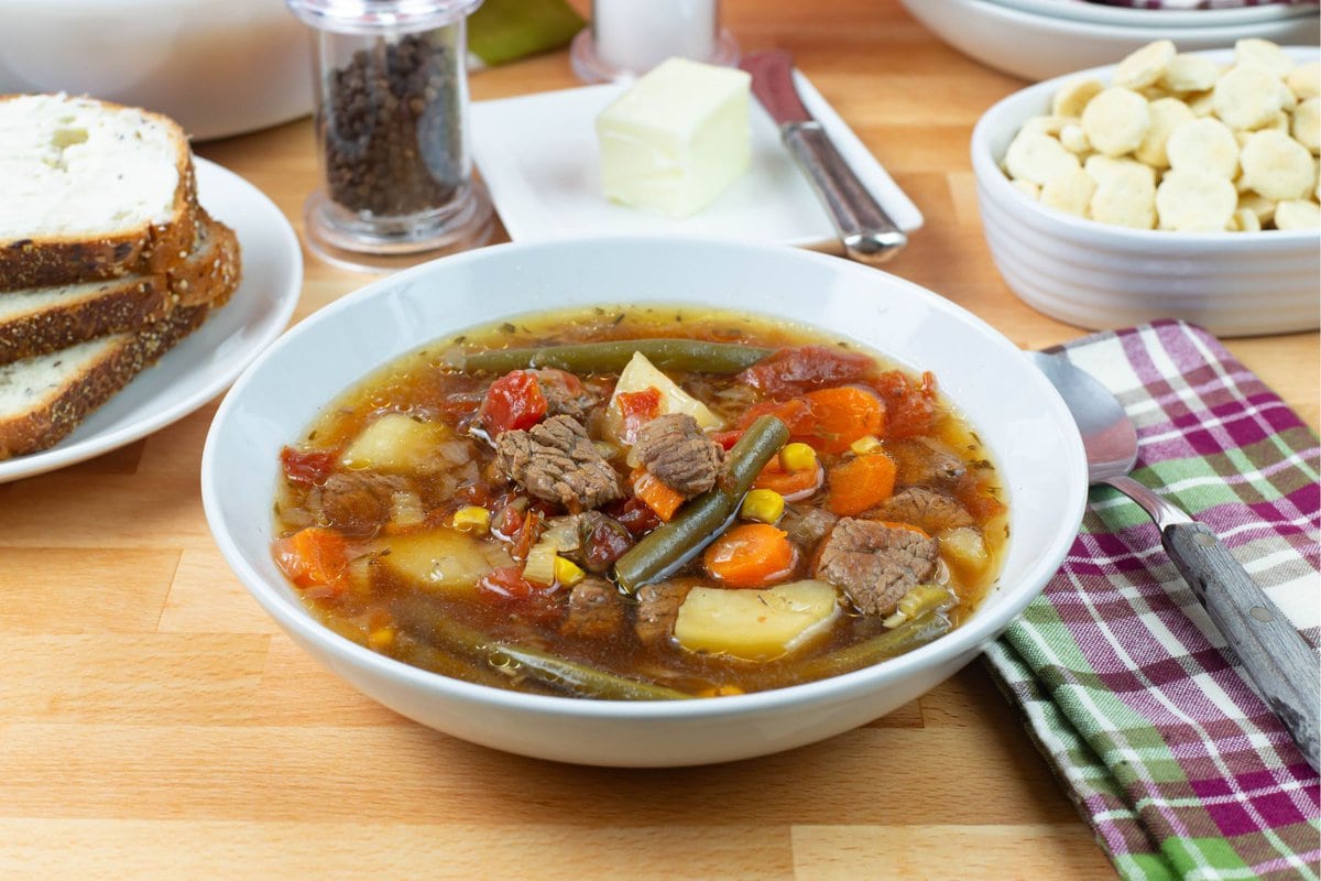 A soup bowl of Beef Vegetable soup with bread and crackers in the background.