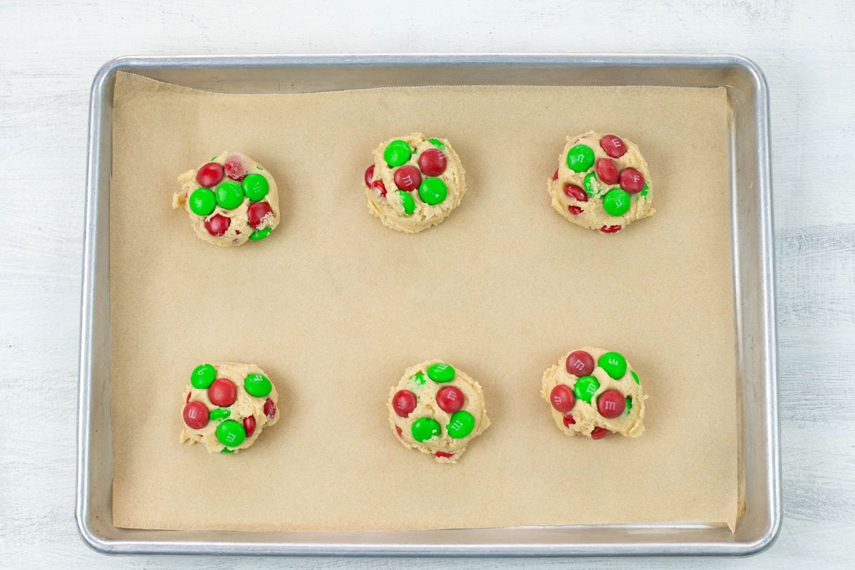 M&M cookie dough balls with red and green m and m's pressed into the dough on a lined cookie sheet.