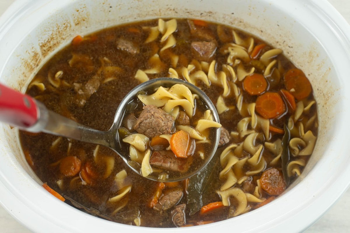 A closeup of a ladle filled with Slow cooker beef and noodle soup.