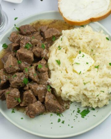 A plate with beef tips and gravy served with mashed potatoes on a dinner plate. Sliced bread in the background.