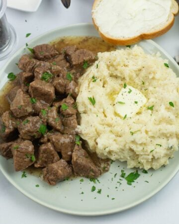 A plate with beef tips and gravy served with mashed potatoes on a dinner plate. Sliced bread in the background.