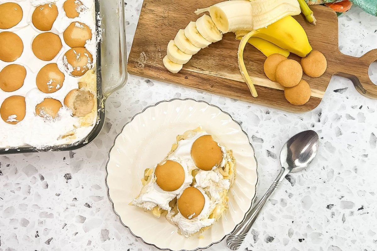 A serving of banana pudding on a plate with a cut banana, and the tray of banana pudding in the background.