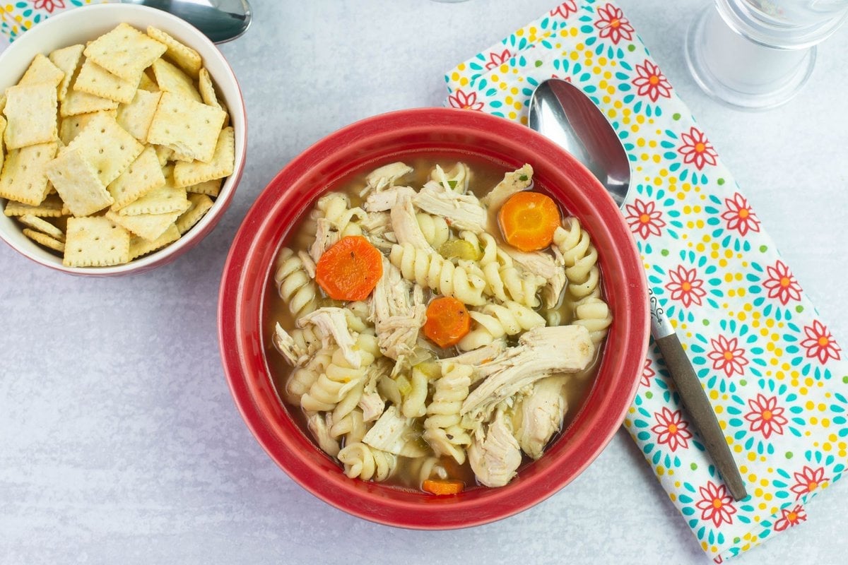 A serving of Spicy chicken noodle soup in a red bowl with a bowl of soup crackers, napkin and spoon beside the bowl.