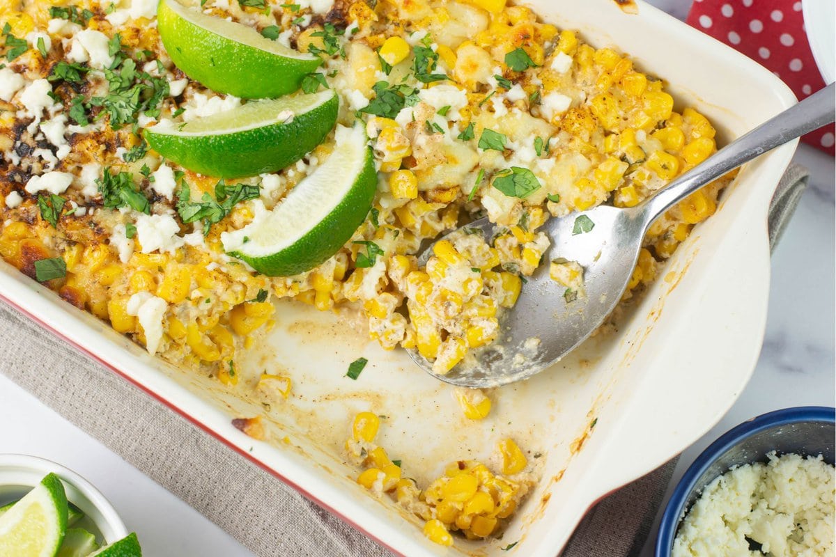 Baking dish with a serving removed of Spicy Creamy Corn side dish topped with lime wedges and cilantro.