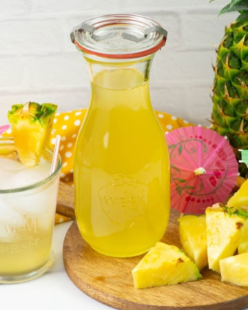Glass decanter of pineapple syrup with a glass filled with Pineapple Soda on a table.