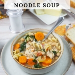 Tall vertical image of a bowl of Tuscan chicken noodle soup on a table with sliced bread in the background.