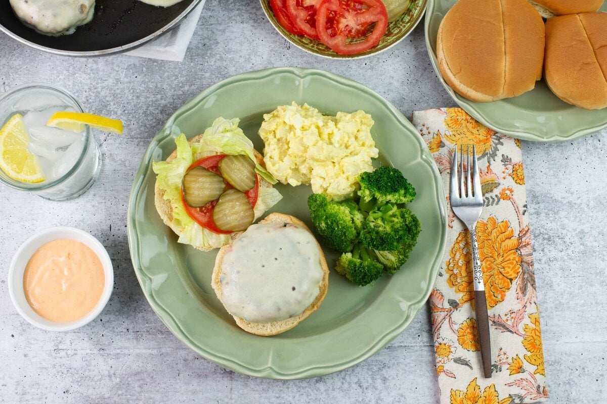 Plated chicken burger with potato salad, broccoli, and toppings.